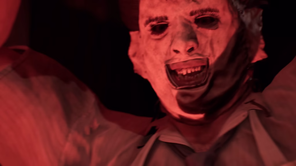 Capturas de tela do jogo The Texas Chain Saw Massacre. Um homem usando uma máscara feita de pele humana (leatherface), com uma expressão facial agressiva e voraz, iluminado por uma luz vermelha intensa em um ambiente escuro.